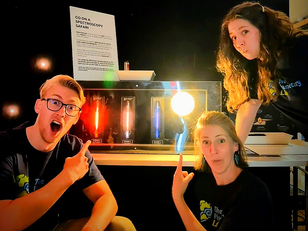 Three cheerful Planetary Lab team members making excited expressions demonstrating the interactive spectroscopy safari exhibit.